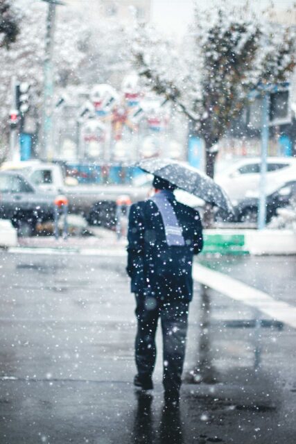 person in black jacket and black pants holding umbrella walking on street during snow